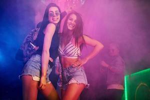 Two beautiful girls dancing in front of young people that having fun in night club with colorful laser lights photo