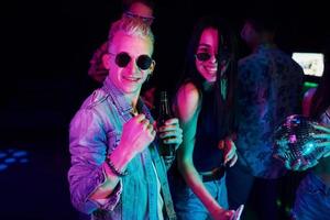 Hipster guy in sunglasses and with bottle of alcohol posing for camera in front of young people that having fun in night club with colorful laser lights photo