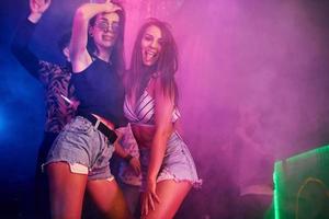 Two beautiful girls dancing in front of young people that having fun in night club with colorful laser lights photo