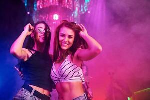 Two beautiful girls dancing in front of young people that having fun in night club with colorful laser lights photo