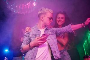 Guy standing with smartphone in front of oung people that having fun in night club with colorful laser lights photo