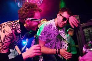 Drunk guy leaning in fence in front of young people that having fun in night club with colorful laser lights photo