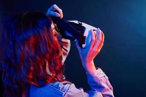 Rear view of girl with curly hair holding virtual reality glasses in red and blue neon in studio photo