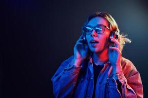 Portrait of young alternative girl in glasses with green hair in red and blue neon light in studio that listening to music in headphones photo