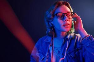 Portrait of young alternative girl in glasses with green hair in red and blue neon light in studio that listening to music in headphones photo