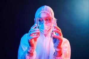 enfermera con máscara y uniforme blanco parada en una habitación iluminada con neón y sosteniendo tubos con muestras