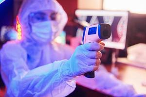 enfermera con máscara y uniforme blanco, sosteniendo un termómetro infrarrojo y sentada en un laboratorio iluminado con neón con computadora y equipo médico en busca de la vacuna contra el coronavirus foto
