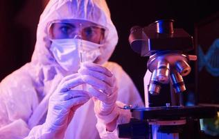 enfermera con máscara y uniforme blanco, sosteniendo una jeringa y sentada en un laboratorio iluminado con neón con computadora y equipo médico en busca de la vacuna contra el coronavirus foto