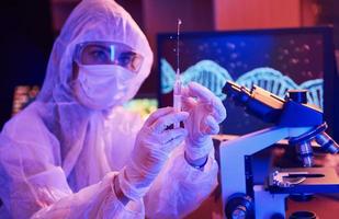 Nurse in mask and white uniform, holding syringe and sitting in neon lighted laboratory with computer and medical equipment searching for Coronavirus vaccine photo