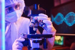 Nurse in mask and white uniform sitting in neon lighted laboratory with computer, microscope and medical equipment searching for Coronavirus vaccine photo