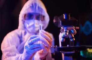 Nurse in mask and white uniform, holding syringe and sitting in neon lighted laboratory with computer and medical equipment searching for Coronavirus vaccine photo