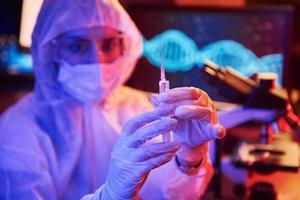 Nurse in mask and white uniform, holding syringe and sitting in neon lighted laboratory with computer and medical equipment searching for Coronavirus vaccine photo