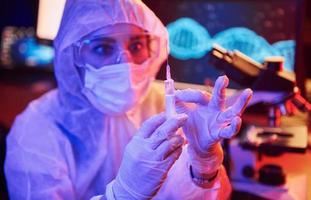 Nurse in mask and white uniform, holding syringe and sitting in neon lighted laboratory with computer and medical equipment searching for Coronavirus vaccine photo