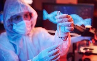 Nurse in mask and white uniform, holding tube with liquid and sitting in neon lighted laboratory with computer and medical equipment searching for Coronavirus vaccine photo