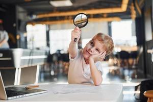 Smart child in casual clothes with laptop on table have fun with magnifying glass photo