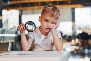 Smart child in casual clothes with laptop on table have fun with magnifying glass photo