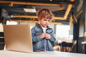 Smart child in casual clothes with laptop on table have fun with magnifying glass photo