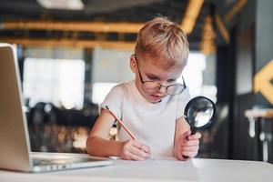 Smart child in casual clothes with laptop on table have fun with magnifying glass photo