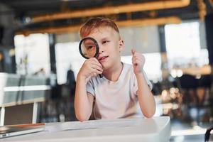 Smart child in casual clothes with laptop on table have fun with magnifying glass photo