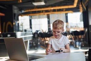 Smart child in casual clothes with laptop on table have fun with magnifying glass photo