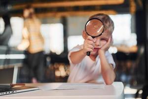 Smart child in casual clothes with laptop on table have fun with magnifying glass photo