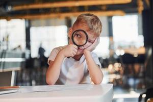 Smart child in casual clothes with laptop on table have fun with magnifying glass photo