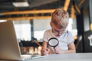 Smart child in casual clothes with laptop on table have fun with magnifying glass photo