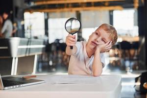 niño inteligente con ropa informal con portátil en la mesa diviértete con lupa foto