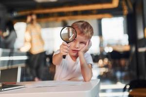 Smart child in casual clothes with laptop on table have fun with magnifying glass photo