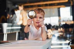 Smart child in casual clothes with laptop on table have fun with magnifying glass photo