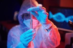 Nurse in mask and white uniform, holding tube with liquid and sitting in neon lighted laboratory with computer and medical equipment searching for Coronavirus vaccine photo