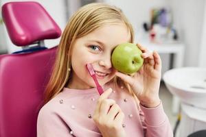 Holding brush and green apple. Little girl visiting dentist in clinic. Conception of stomatology photo