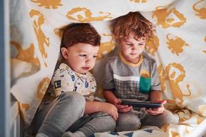 Two little boys resting and have fun indoors under blanket together with smartphone photo
