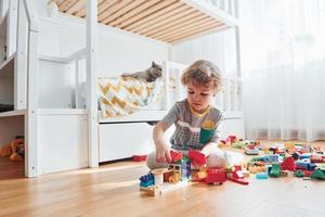 Cute little boy sitting and have fun indoors in the bedroom with plastic construction set. Cat at background photo