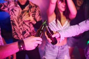 Celebrating and knocking bottles with alcohol. Young people is having fun in night club with colorful laser lights photo