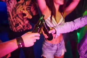 Celebrating and knocking bottles with alcohol. Young people is having fun in night club with colorful laser lights photo