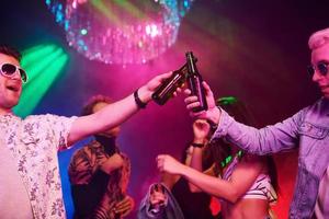 Celebrating and knocking bottles with alcohol. Young people is having fun in night club with colorful laser lights photo