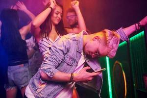 Guy standing with smartphone in front of oung people that having fun in night club with colorful laser lights photo