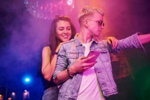 Guy standing with smartphone in front of oung people that having fun in night club with colorful laser lights photo