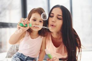 Young mother with her daughter blowing bubbles together in bedroom photo