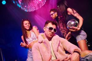 Elegant man in sunglasses in the midle feels like a boss. Young people is having fun in night club with colorful laser lights photo