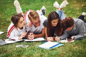 Writing on paper. front view. Group of young students in casual clothes on green grass at daytime photo