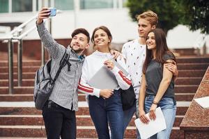 de pie y tomando selfie. grupo de jóvenes estudiantes con ropa informal cerca de la universidad durante el día foto