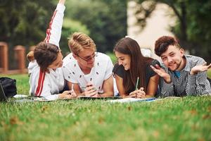 Writing on paper. front view. Group of young students in casual clothes on green grass at daytime photo