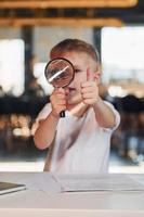 Smart child in casual clothes with laptop on table have fun with magnifying glass photo