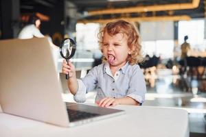 Smart child in casual clothes with laptop on table have fun with magnifying glass photo