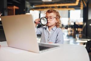 niño inteligente con ropa informal con portátil en la mesa diviértete con lupa foto