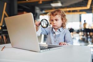 Smart child in casual clothes with laptop on table have fun with magnifying glass photo