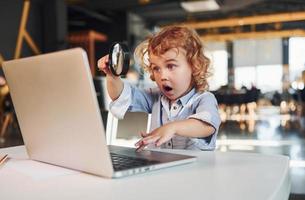 Smart child in casual clothes with laptop on table have fun with magnifying glass photo