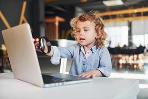 Smart child in casual clothes with laptop on table have fun with magnifying glass photo